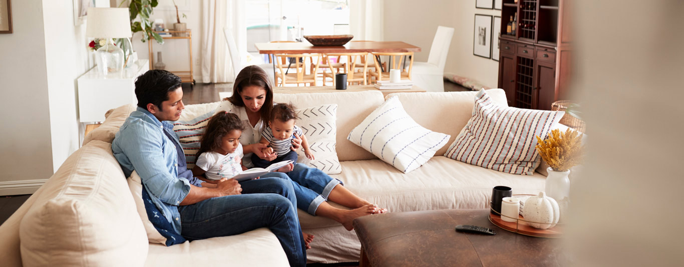 iStock-1094435430-1366×534-M Family sitting on the sofa looking at a book together